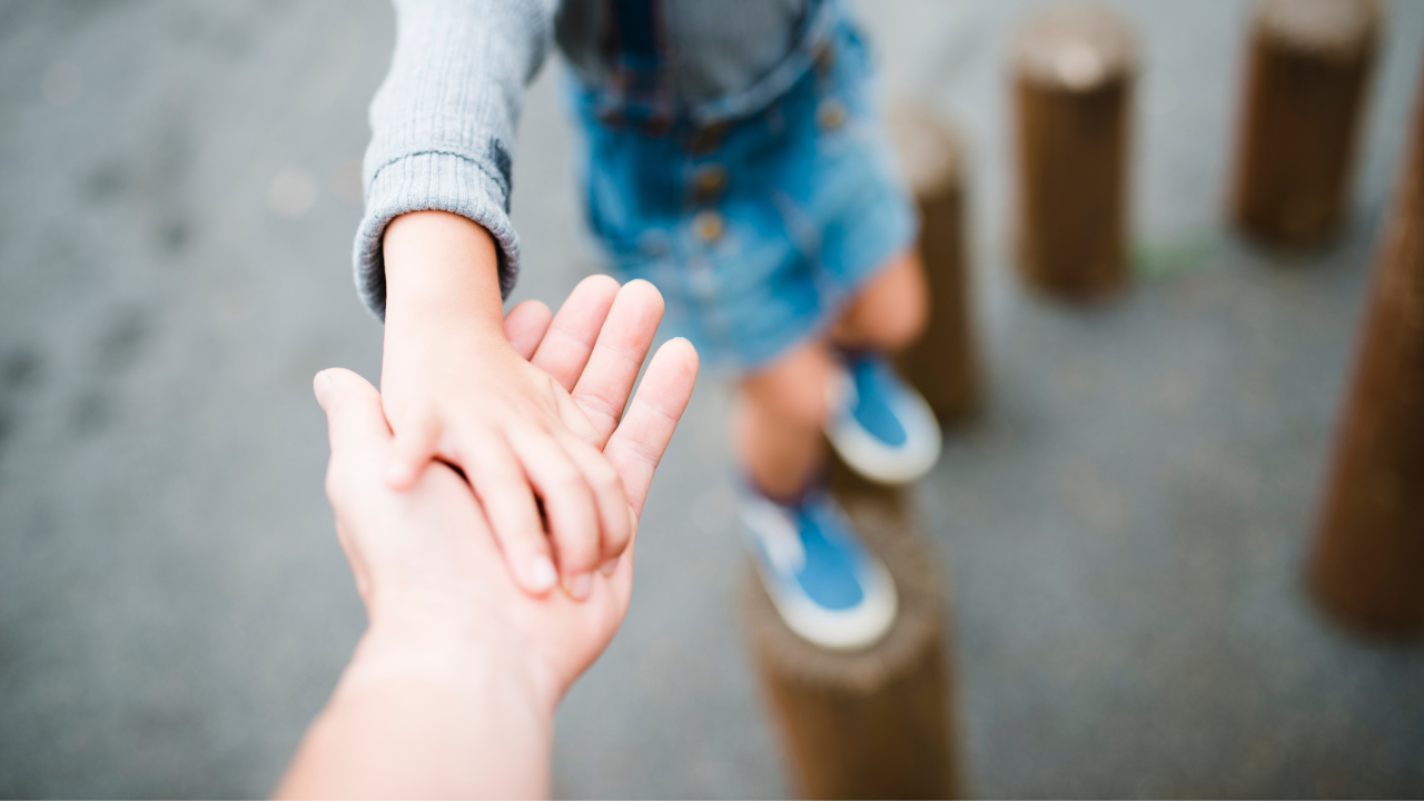 An adult’s hand holds a child’s hand as the child balances on wooden posts.